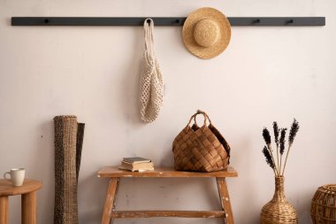 The stylish composition of cosy entryway with wooden bench, coffee table, basket, hanger and personal accessories. Beige wall. Home decor. Template. 
