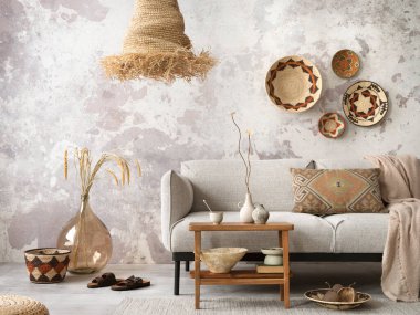 The stylish ethnic composition at living room interior with grey sofa, colorful wicker, straw lamp, bench and elegant personal accessories. Grey concrete wall. Cozy apartment. Home decor. 