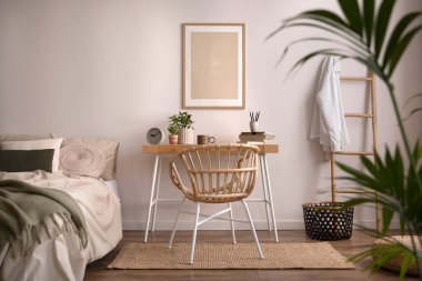 Stylish composition of cozy bedroom with mock up, beige bedding, plants, armchair and wooden desk. Mock up poster with wooden frame. Home decor. Template. 