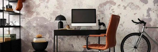 Concrete interior of home office with black desk, brown armchair, computer screen, office accessories and lamp. Rack with personal accessories. Home decor. Template.