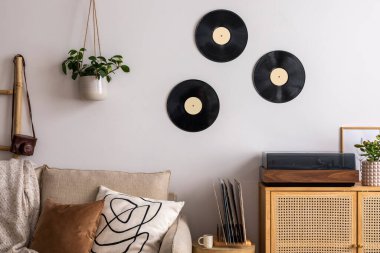 The stylish composition of living room interior with beige sofa, pillow, plaid, vinyl record, rattan commode and personal accessories. Beige wall. Template. 