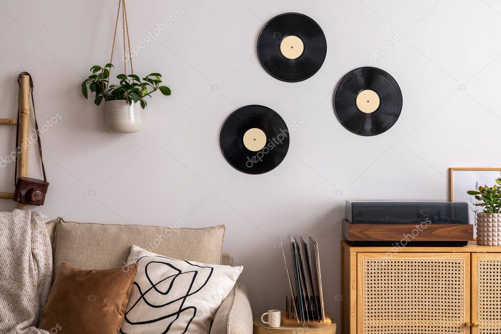 The stylish composition of living room interior with beige sofa, pillow, plaid, vinyl record, rattan commode and personal accessories. Beige wall. Template. 
