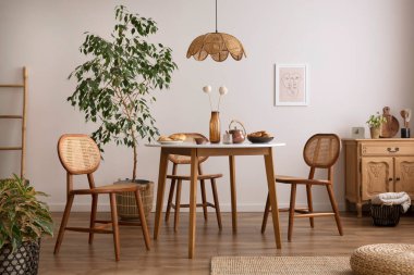 The stylish dining room with round table, rattan chair, wooden commode, poster and kitchen accessories. Beige wall with mock up poster. Home decor. Template. 