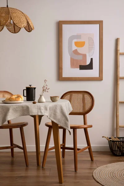 The stylish dining room with round table, rattan chair, dog on the carpet,  poster and kitchen accessories. Beige wall with mock up poster. Home decor.  Template. Stock Photo