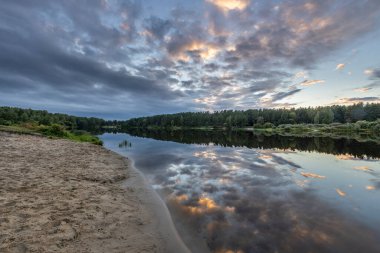 Akşamları nehir kıyısında yaz manzarası. Dramatik bulutlar suya yansıyor. Gün batımından sonra manzarada yoğun renkler.