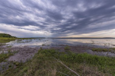 Baharın başlarında göl manzarası. Dramatik gökyüzü göle yansıyor. Ön planda genç Mayıs yeşil çimenleri.