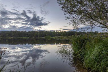 Akşamları nehir kıyısında yaz manzarası. Dramatik bulutlar suya yansıyor. Gün batımından sonra manzarada yoğun renkler.