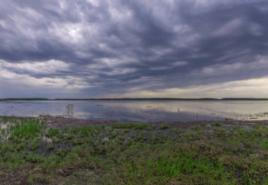 Baharın başlarında göl manzarası. Dramatik gökyüzü göle yansıyor. Ön planda genç Mayıs yeşil çimenleri.
