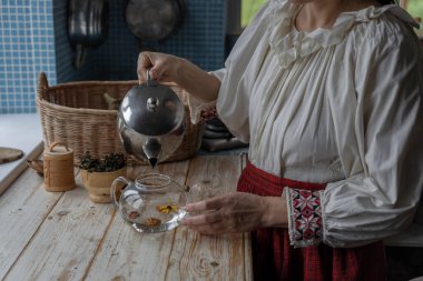 Halk kostümü giymiş bir kız bitki ve çiçeklerden çay yapar. Mutfakta bir kadın kendi çayını hazırlıyor..