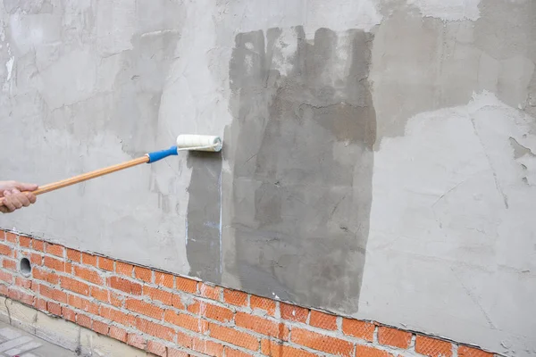 A worker applies the primer to the wall with a long-handled roller. A man\'s hand rolls a primer onto a gray wall. Wet footprint on a plastered wall. House facade renovation concept.