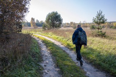 Bir turist sabahın erken saatlerinde köy yolunda yürür. Sırt çantalı bir adam güneş ışığıyla aydınlatılmış bir tarlanın arka planında.
