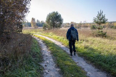 Bir turist sabahın erken saatlerinde köy yolunda yürür. Sırt çantalı bir adam güneş ışığıyla aydınlatılmış bir tarlanın arka planında.