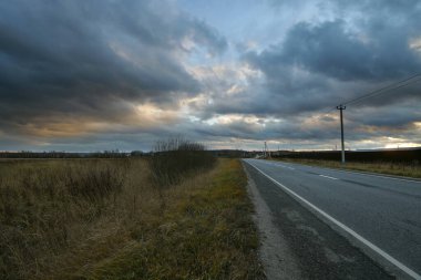 Ufkun ötesine giden bir yolun olduğu akşam manzarası. Sonbahar, bulutlu dramatik gökyüzü. Akşam ışığında rengarenk gökyüzü. Kırsal alan.