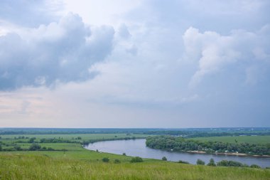 Önünde çayır otları olan yaz manzarası. Yağmurdan önce bulutlu karanlık bir gökyüzü. Parlak yeşil çimenler. Arkaplanda geniş bir nehir.