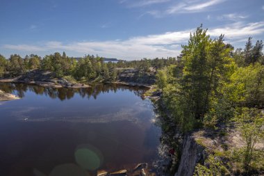Göl üzerinde taşlarla kaplı bir orman manzarası. Gölde güneşli bir gün. Suyun içindeki gökyüzünün yansıması. Taş üstünde çam. Kuzeyin doğası.. 