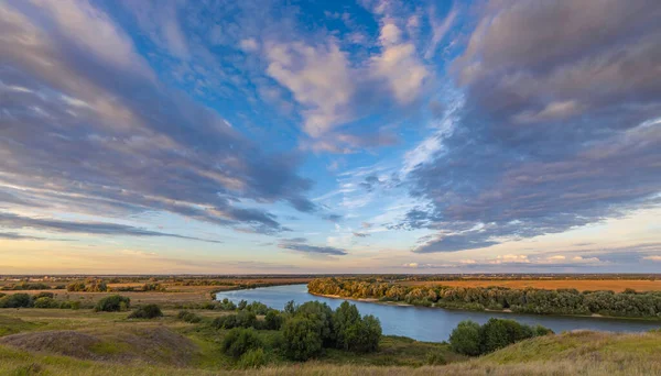Nehirli güneşli yaz manzarası. Güzel panoramik manzara. Yeşil tepeler, tarlalar ve çayırlar. Ufkun ötesindeki dramatik gökyüzü. Sakin ol. Ilık güneş ışığı. Mavi gökyüzünde renkli bulutlar.