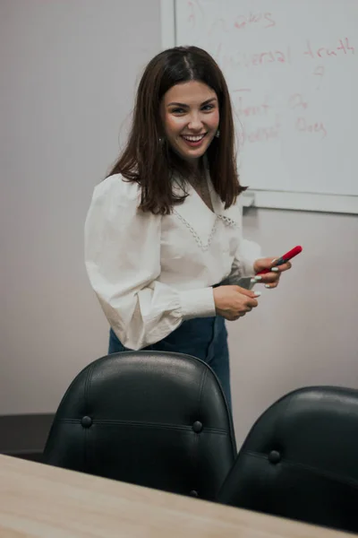 Young female english language teacher with a red marker in a white blouse standing in front of a white board and smiling