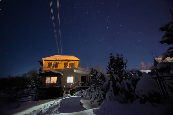 Nachtaufnahme eines Backsteinhauses mit beleuchteten Fenstern und einem Sternenhimmel auf winterlichem Hintergrund. Konzept von Frieden, Magie, Idylle, Heimat, Träumen. — Stockfoto