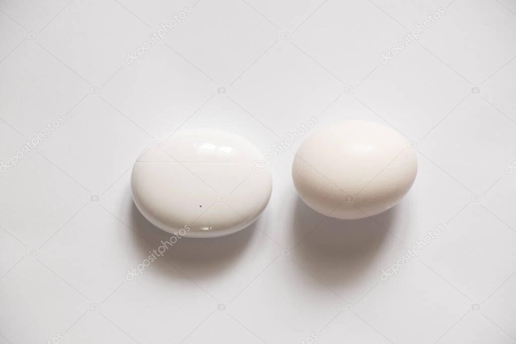 charging case with wireless headphones in the shape of an oval lies and a chicken white egg lie on an isolated background, wireless headphones, a comparison of the shape