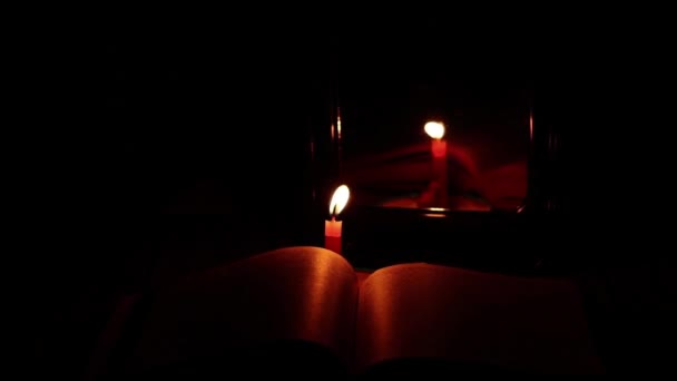 聖書と炎のろうそくが鏡の近くのスツールや祈り宗教の上の暗闇の中で — ストック動画