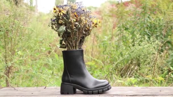 靴の内側に野生の花の花束と木製のテーブルの上に厚い靴底を持つ女性の高い黒いブーツ 女性の冬の靴 履物とファッション — ストック動画