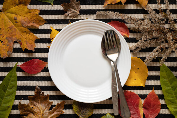 Dinner plate stands on a striped black and white background in autumn yellow-red leaves, autumn table decor, cutlery, table decorations