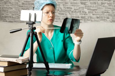 Tıbbi üniformalı bir doktor görüntülü konuşma yoluyla online danışmanlık yapıyor ve ellerinde bir röntgen tutuyor, internette bir tıp konferansı.