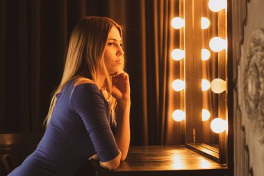 Bir kız karanlık bir odada ampullerle aynanın yanında oturuyor, bir kız stüdyoda aynanın yanında.