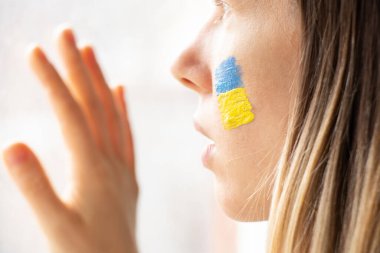 Yüzünde Ukrayna bayrağı olan Ukraynalı bir kadın camdan dışarı bakıyor, vatansever, Ukrayna 'da barış, sıkıyönetim, pencerenin dışındaki savaşı görüyorum, 2022 savaşını durduruyorum.