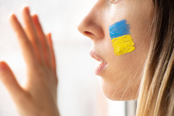 A Ukrainian woman with the national flag of Ukraine yellow-blue on her face looks out the window, patriot, peace in Ukraine, martial law, I see the war outside the window, stop war 2022