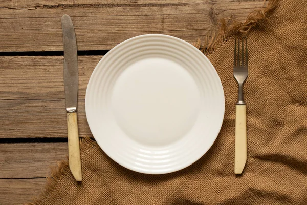 1枚の白いプレートとナイフ付きのフォークは 自宅のキッチンで古い木製のテーブルの上にあります — ストック写真