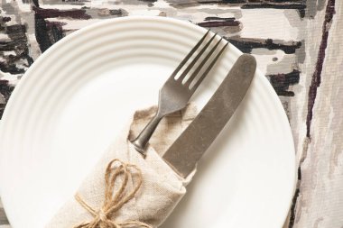 Eskimiş çatal ve bıçak peçeteye sarılı gri bir arka planda beyaz bir tabakta yatıyor. Kelime servisi, çatal bıçak takımı.