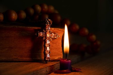 A bible cross and a wooden rosary lie on the table next to candles burning in the dark at home, prayer and faith, god and religion