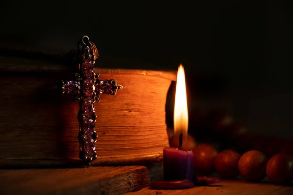 聖書の十字架と木製のロザリオは 祈りと信仰 神と宗教で暗闇の中で燃えるろうそくの横にテーブルの上に横たわっています — ストック写真