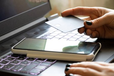 Bir kız elinde siyah bir dizüstü bilgisayarın yanında bir kredi kartı ve pencerenin yanındaki ofiste bir telefon tutuyor, online ödeme, internet kartıyla ödeme.