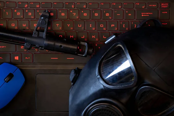La ametralladora militar y el ratón de la computadora y la máscara de gas negro se encuentran en el teclado del ordenador portátil con luz de fondo roja, ataque de hacker, cibernético —  Fotos de Stock