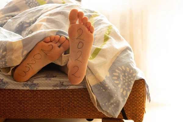 Jambes d'une fille sur un lit à la maison sous une couverture, 2022 et 2023 sont écrits sur ses jambes, le début d'une nouvelle année, bonne année — Photo