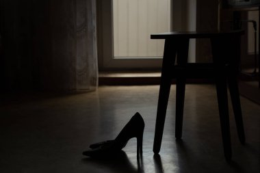 Kadınların yüksek topuklu ayakkabıları evin zemininde duruyor. Karanlıkta tahta bir sandalyenin yanında, şık kadın ayakkabıları, topuklu ayakkabılar.