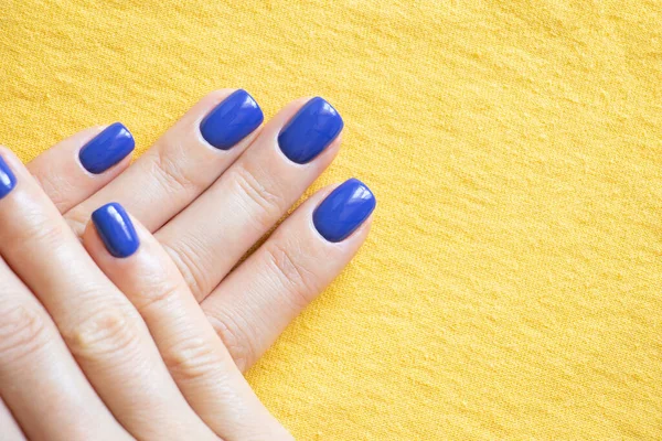 Manicure and blue polish on the girl\'s finger on a yellow background, yellow-blue color of the flag of Ukraine, gel polish on the girl\'s nails, manicure