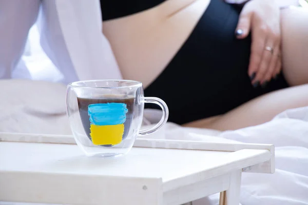 A pregnant girl is sitting near a cup of coffee on which the flag of Ukraine is drawn, a new life during the war in the country, the victory of Ukraine, the flag of Ukraine on the cup
