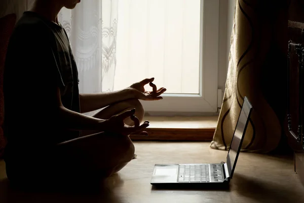 A girl is doing yoga near a laptop on the floor of a house near a window in the dark, an online video lesson on meditation, yoga and miditatsi