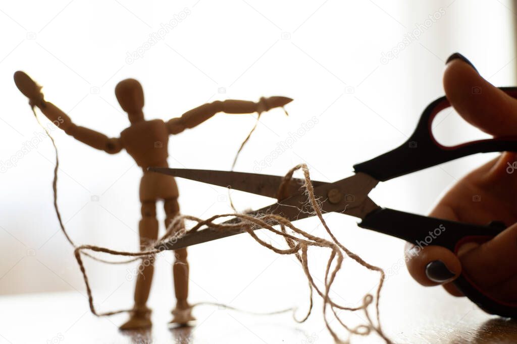 Wooden puppet tied with ropes to the puppeteer and scissors cut the rope, manipulation of people and freedom, strength and freedom over power
