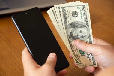 Masada oturan bir adamın elinde dolar ve telefon, online ödeme, para ve finans.