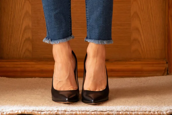Women\'s legs in jeans and black high-heeled shoes on the floor of the house on the carpet near the door, women\'s shoes with heels, legs in shoes