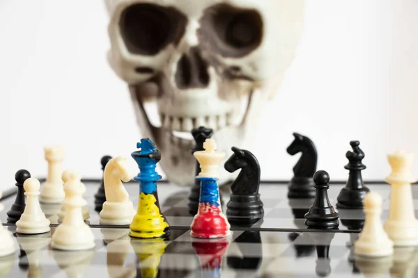 Chess with a painted flag of Russia and Ukraine on a chessboard and a head of a skeleton in the background, a death game between the two countries of Ukraine and Russia, war