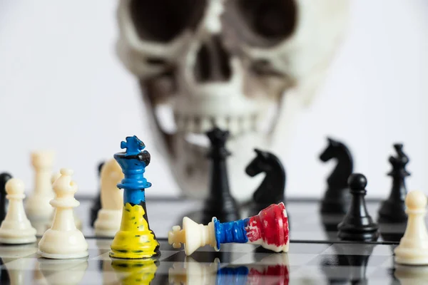 Chess with a painted flag of Russia and Ukraine on a chessboard and a head of a skeleton in the background, a death game between the two countries of Ukraine and Russia, war