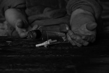 Sigara bir bardak ve boş bir şişe ve yanmış siyah bir masanın üzerinde yaşlı bir kadının elleri, sigara ve alkolizm, kötü alışkanlıklar.