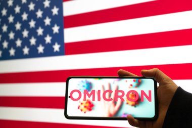 hand holding mobile phone with omicron COVID 19 variant strain in front of flag of USA showing news app notification clipart