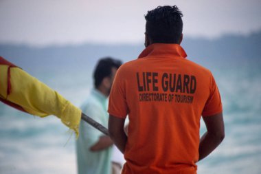 Havelock Swaraj dweep and man Nicobar Adaları 'nda arka planda turistlerle sahil boyunca bayrak taşıyan turuncu tişörtlü cankurtaran.