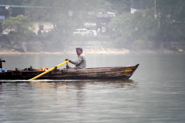 Nanital, Uttarakhand, India - circa 2022 : local man in a colorful traditional cap rowing an elegant row boat with seats for tourists to travel in the foggy lake of Naini jheel in this famous tourist
